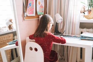 Pretty school girl with long hair in red dress draws with pencil at table in children's room at home