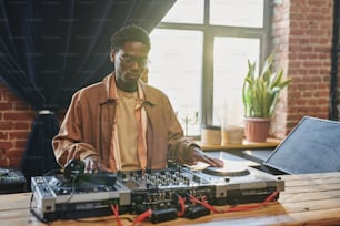 Junger ernsthafter schwarzer Mann, der auf das DJ-Board schaut, während er Musikgeräte anpasst und Plattenspieler berührt, bevor er auftritt