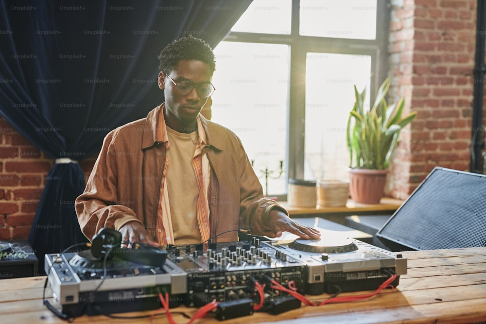 Young serious black man looking at dj board while adjustic musical equipment and touching turntables before performing