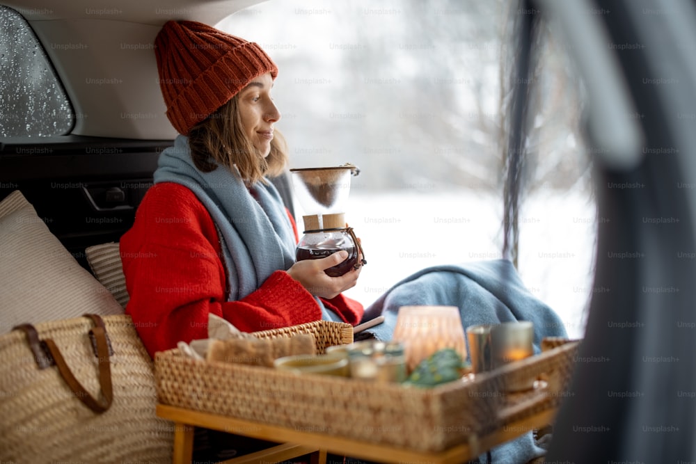 Woman preparing coffee using glass coffeemaker pour over coffee maker in car trunk, traveling by car during winter holidays. High quality photo