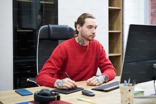 Young designer sitting at desk in front of computer monitor and using graphic tablet to do his work