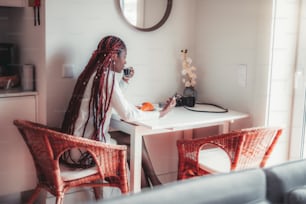 Una giovane donna nera sensuale abbagliante sta facendo colazione nella cucina di casa sua, bevendo un espresso e leggendo il feed di notizie sullo schermo del suo smartphone mentre è seduta su una poltrona di vimini