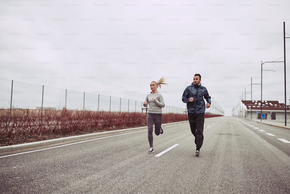 Casal jovem diversificado em roupas esportivas olhando focado enquanto correm juntos ao longo de uma estrada no país