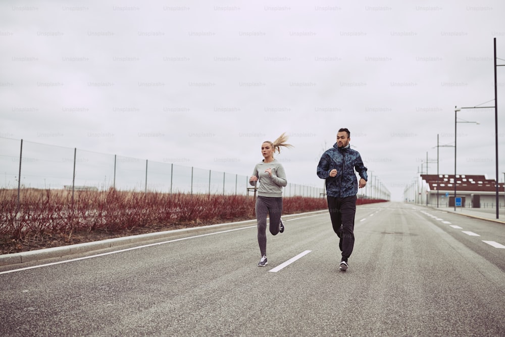 Pareja joven diversa en ropa deportiva que se ve concentrada mientras corren juntos a lo largo de una carretera en el campo