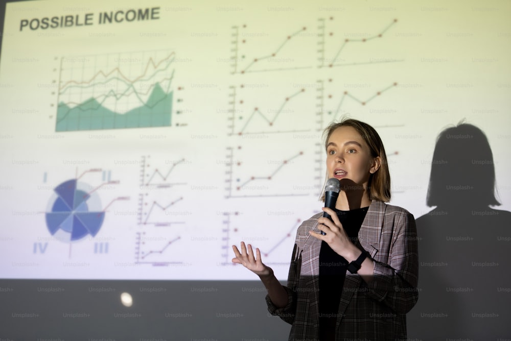 Young contemporary broker of financial analyst standing against large screen with graph during speech in front of audience