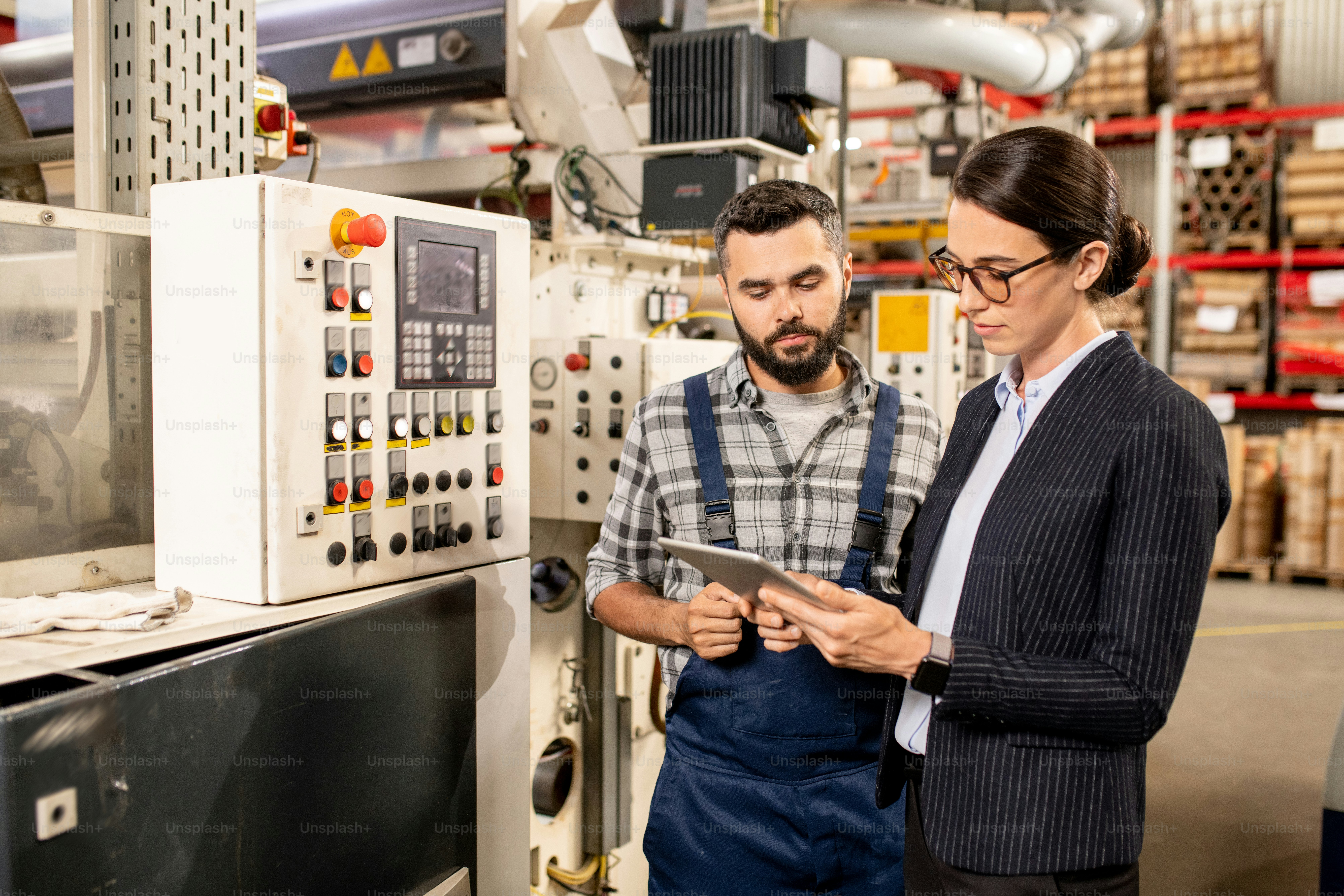 Junge Geschäftspartnerin in Formalwear scrollt im Tablet auf der Suche nach technischen Daten mit Ingenieur in der Nähe