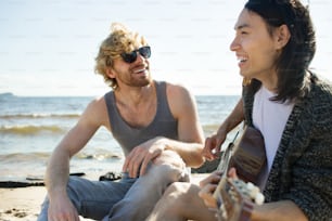 Two young men smiling and playing guitar while sitting on beach during party.