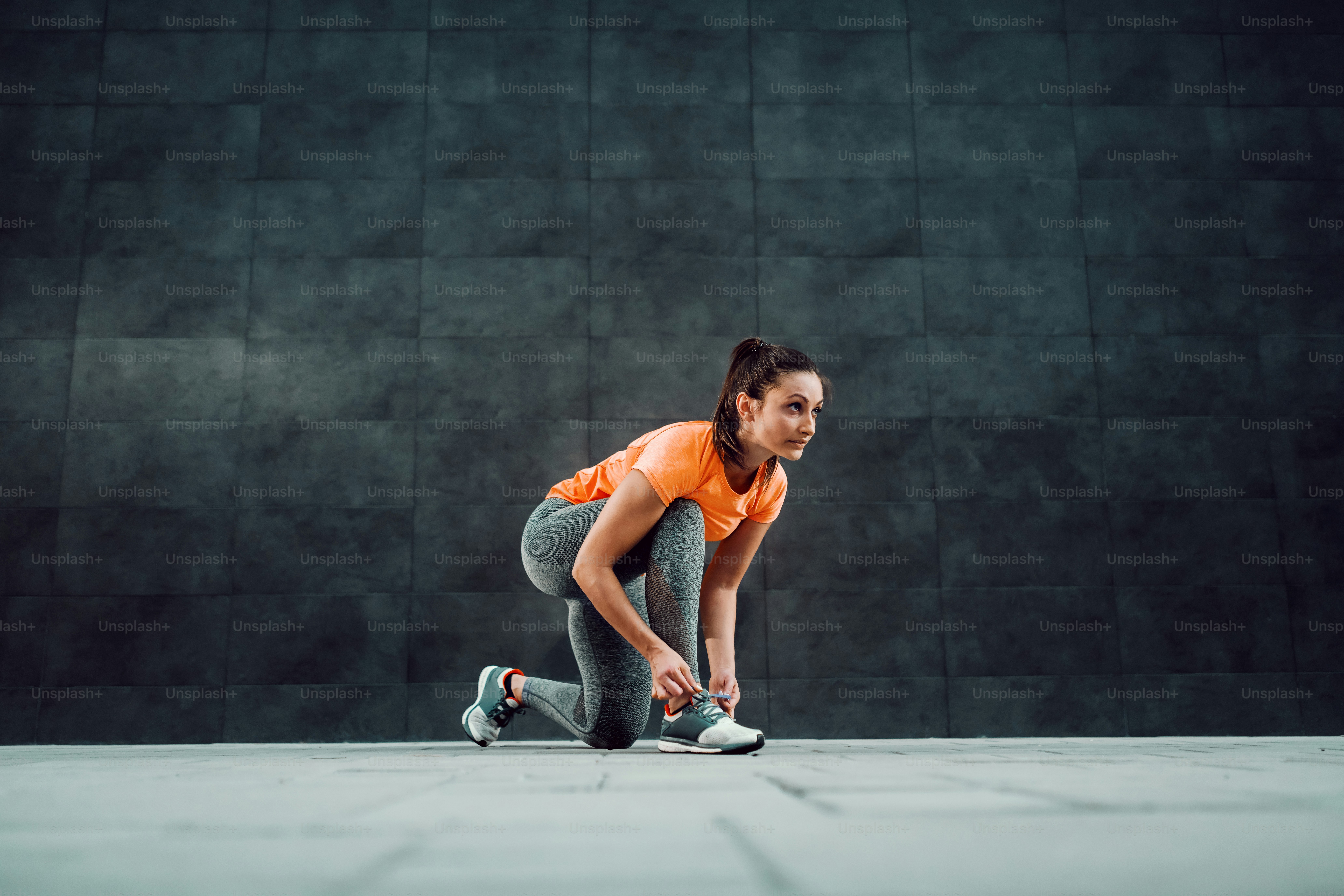Ponte una atractiva morena caucásica en ropa deportiva y con cola de caballo, arrodillándote y atándote los cordones de los zapatos antes de correr.