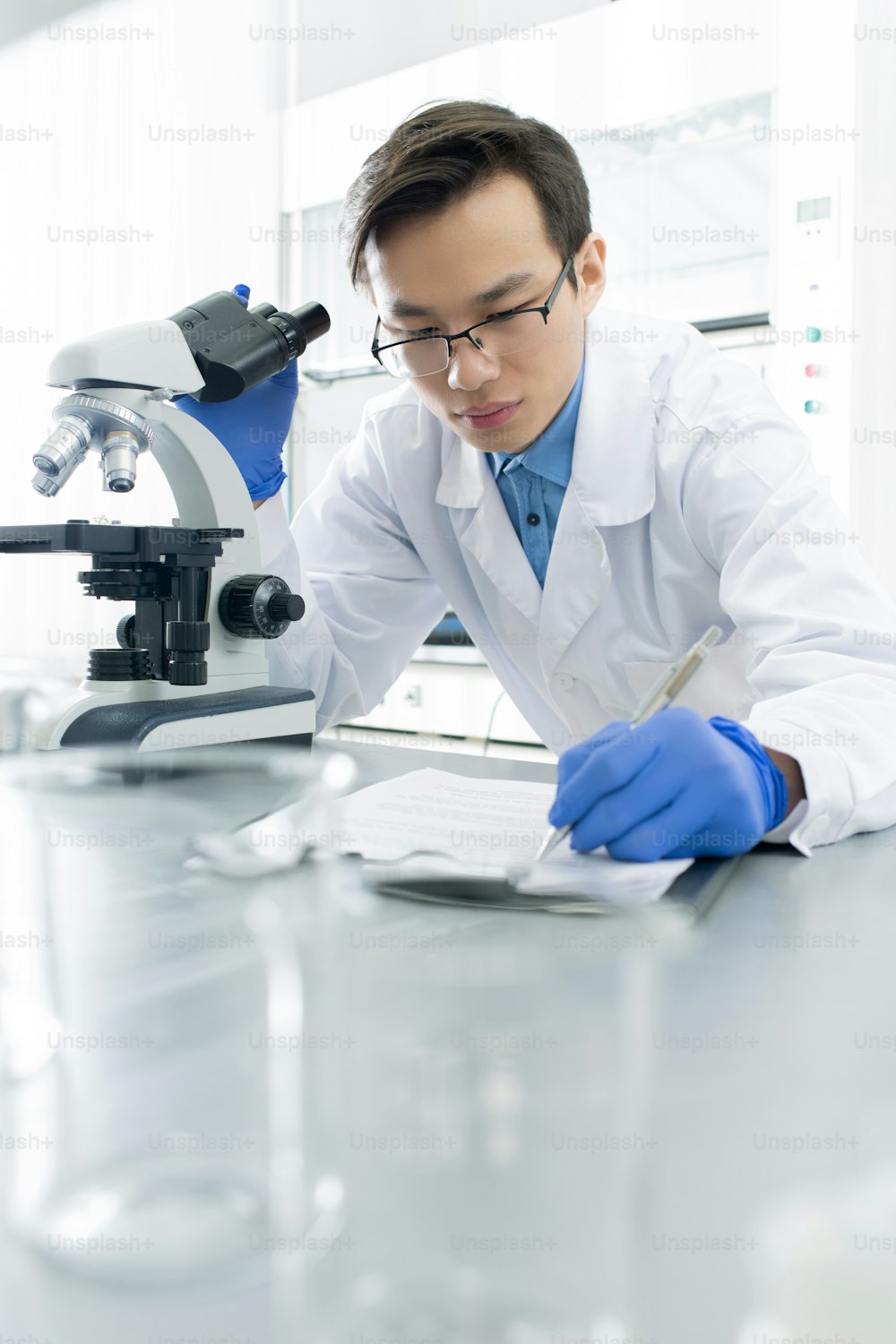 장갑을 끼고 흰 코트를 입은 중국 남성 실험실 직원이 책상을 구부리고 현미경으로 새로운 바이러스를 연구하면서 메모장에 메모를 하고 있다