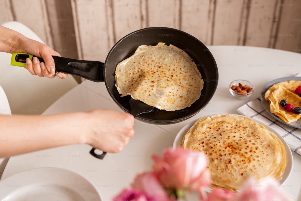 Hands of young woman holding frying pan over kitchen table while taking hot appetizing pancake to put it on top of other crepes on plate