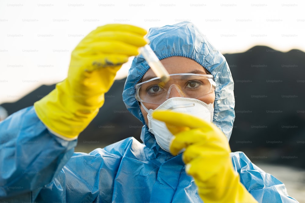 Young female researcher in protective workwear looking at flask with sample of toxic soil while making investigation in dangerous area