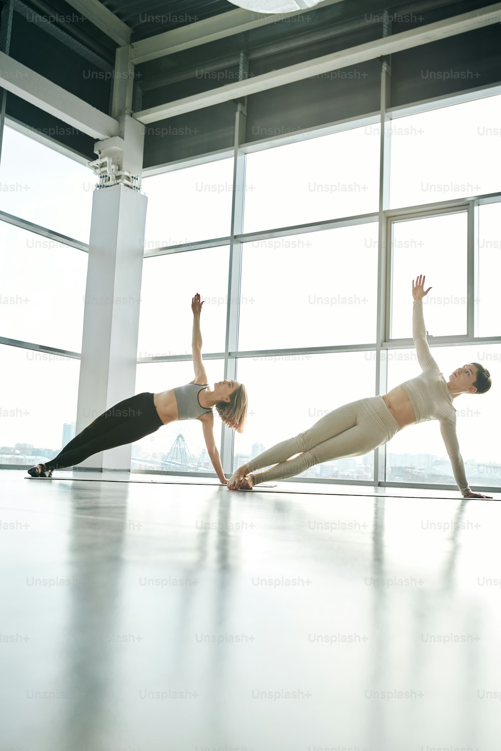 Two fit girls in sportswear exercising with stretched arms on mats while training against large windows at leisure center