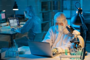 Young serious female virologist in protective workwear typing on laptop keyboard while sitting by desk during scientific research