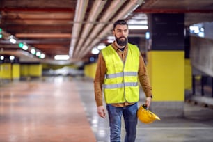 ベストを着たハンサムな髭を生やした労働者の4分の3の長さが安全ヘルメットを持って職場に戻る。建設中の地下駐車場。