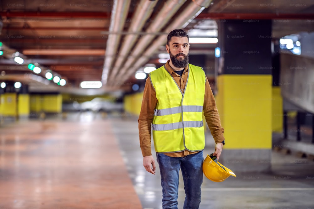 Three quarter length of handsome bearded worker in vest holding his safety helmet and going back to his workplace. Underground garage in construction process interior.