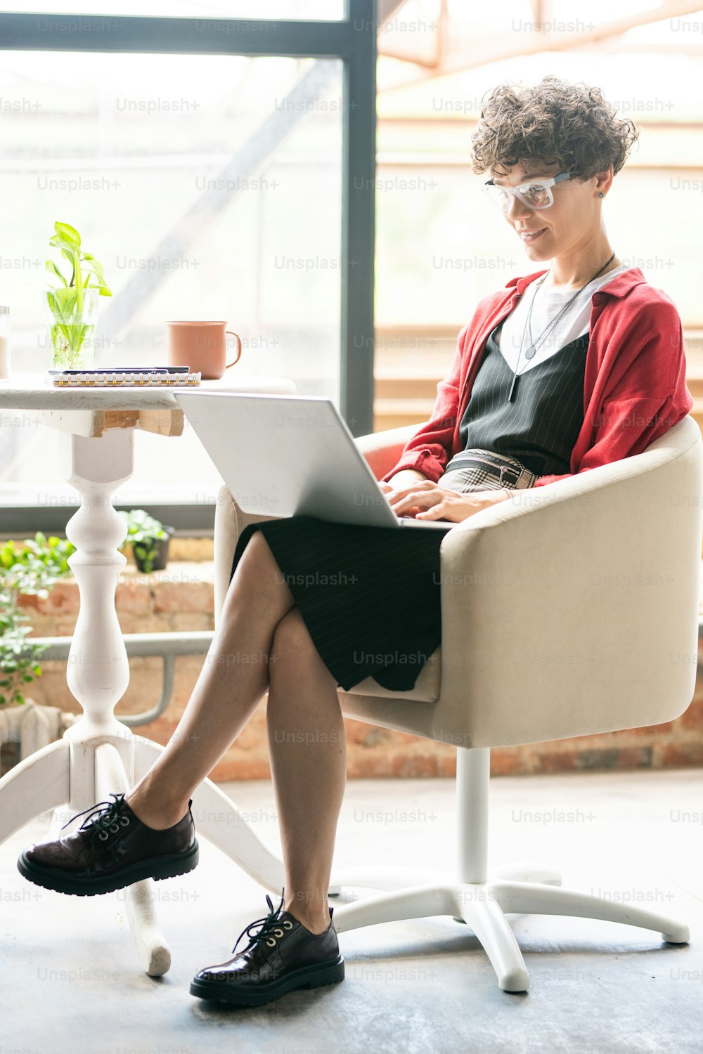 Joven elegante mujer de negocios que hace el trabajo de la computadora mientras está sentada en el sillón con la computadora portátil en el frente dentro de la oficina