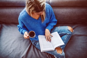 스웨터를 입은 금발 백인 젊은 여성이 거실 소파에 앉아 커피를 마시고 책을 읽고 있다.