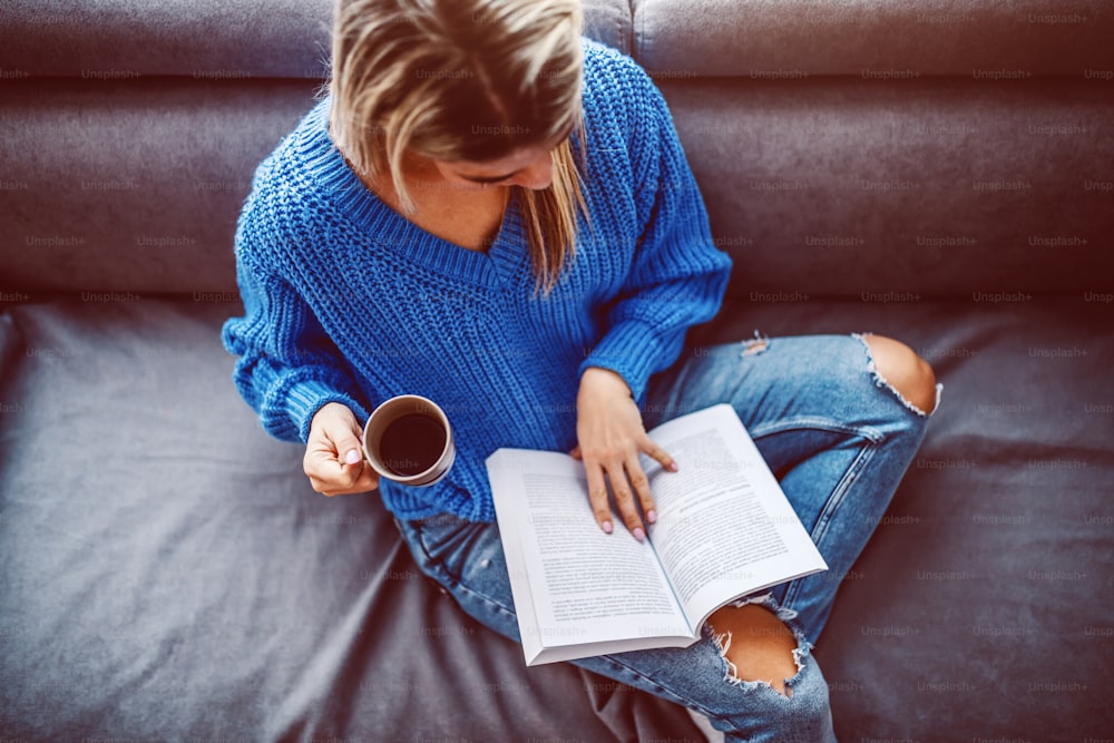 Vue de dessus d'une jeune femme caucasienne blonde en pull assis sur un canapé dans le salon, buvant du café et lisant un livre.