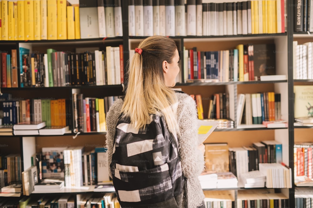 Rear view of young blonde college student girl with a backpack choosing a book from the library bookshelf.
