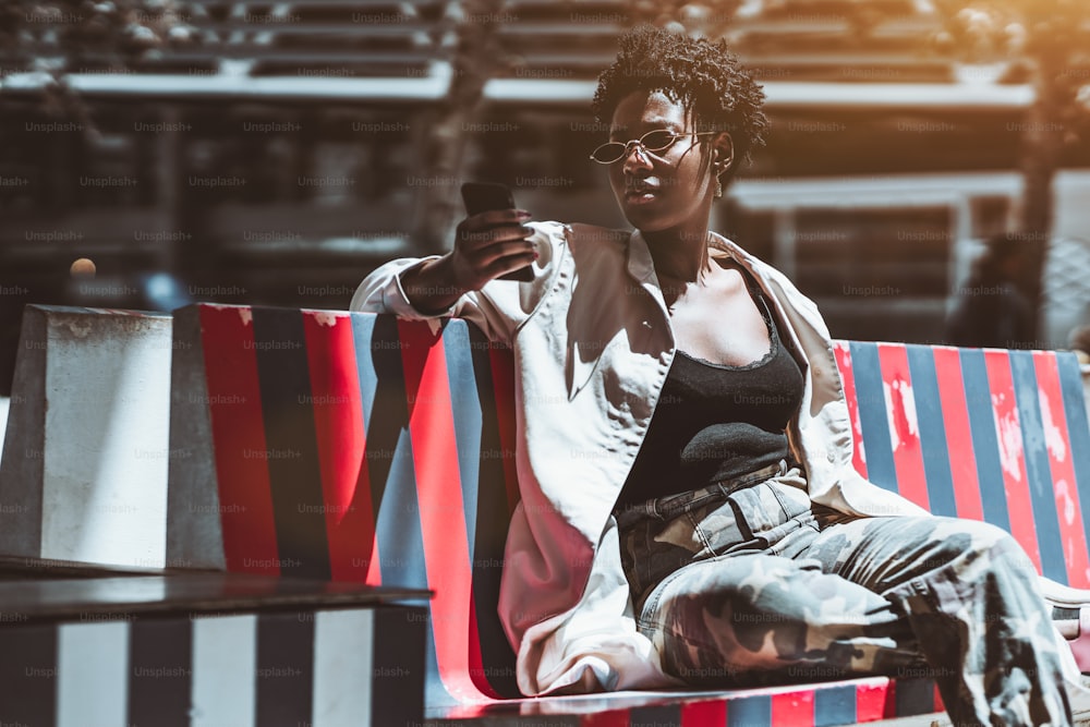 An elegant young African female is using her cellphone while sitting on a striped street bench; a fancy black girl in sunglasses and a white trench is phoning outdoors on a sunny day
