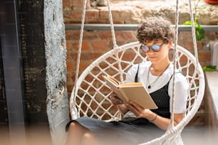 Contemporary young woman in casualwear and eyeglasses reading novel or other literature at leisure while relaxing at home