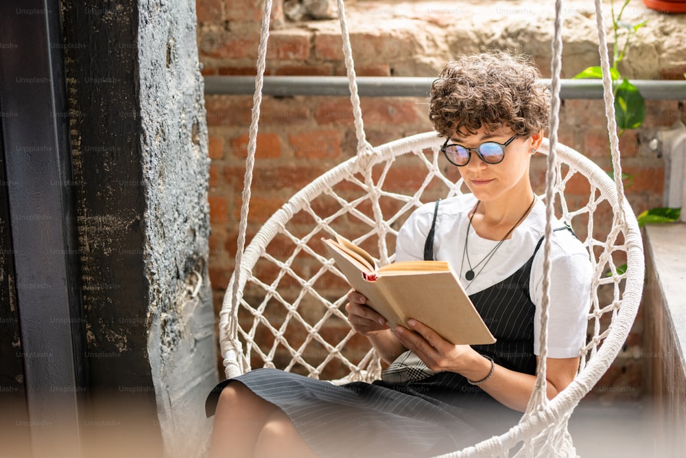 Mujer joven contemporánea en ropa casual y anteojos leyendo novela u otra literatura en el ocio mientras se relaja en casa