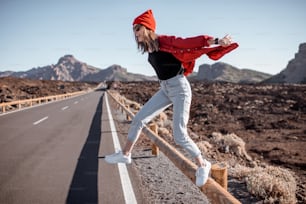 Lifestyle-Porträt einer jungen, stilvoll in Rot gekleideten Frau, die auf der Straße springt und an einem sonnigen Tag durch das Vulkantal reist. Sorgloses Lifestyle- und Reisekonzept