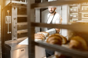 Baker carrying shovel with fresh baked breads standing near the professional oven in the bakery. View behind the bread shelve