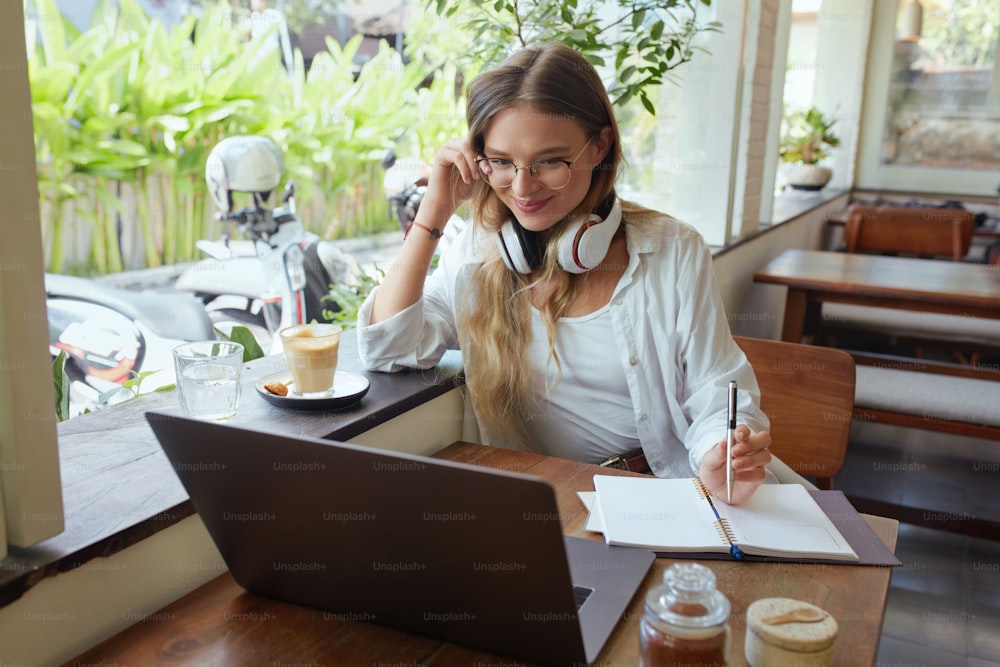 Work. Woman With Laptop And Headphones At Coffee Shop. Happy Stylish Girl In Glasses Looking At Screen And Going To Write In Planner Book. Digital Technologies For Online Working Or Studying At Cafe.