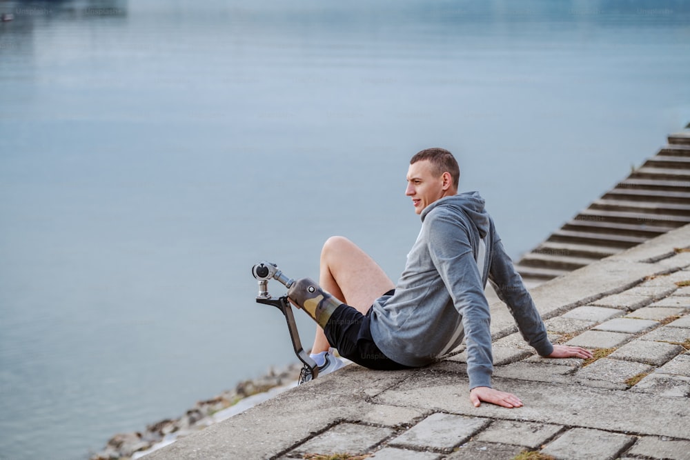 Side view of caucasian handicapped sportsman in sportswear and with artificial leg sitting on quay and looking at river.