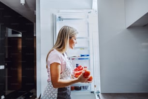 Sorridente dona de casa loira caucasiana no avental tirando legumes da geladeira. A mulher quer preparar o jantar para sua família.