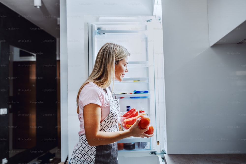 Sorridente dona de casa loira caucasiana no avental tirando legumes da geladeira. A mulher quer preparar o jantar para sua família.