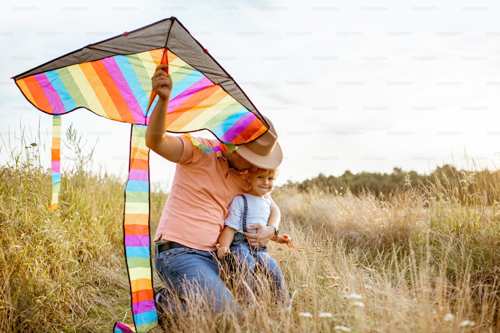 Retrato de um pai e filho felizes segurando pipa de ar colorida enquanto sentados juntos no campo durante o pôr do sol
