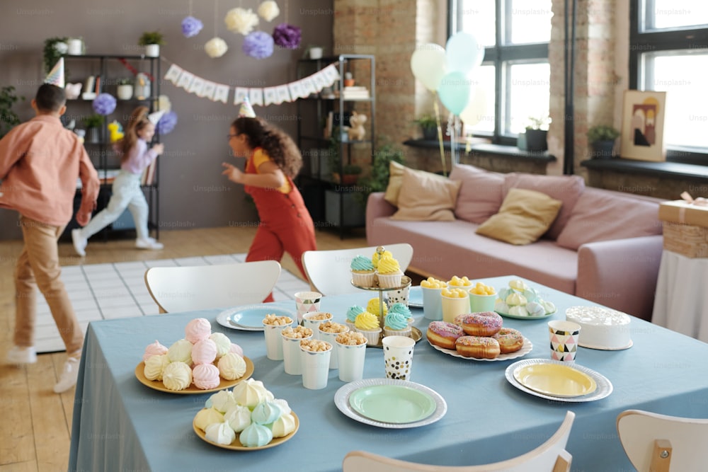 Image of table with desserts and sweet food with group of children running and having fun together in background at party
