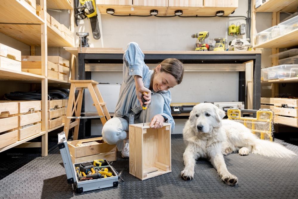 Young handywoman makes a wooden boxes, sitting on the floor with her cute dog in the well equipped workshop at home
