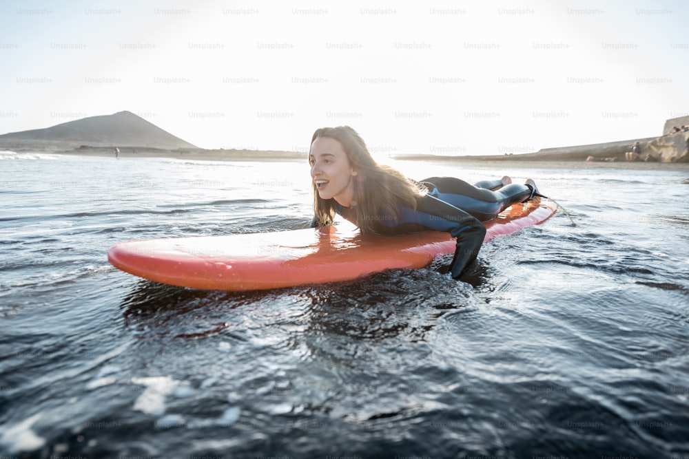 잠수복을 입은 젊은 여자는 서핑 보드에서 물의 흐름을 잡고, 일몰 동안 물결 모양의 바다에서 서핑을 합니다. 수상 스포츠와 활동적인 라이프 스타일 개념