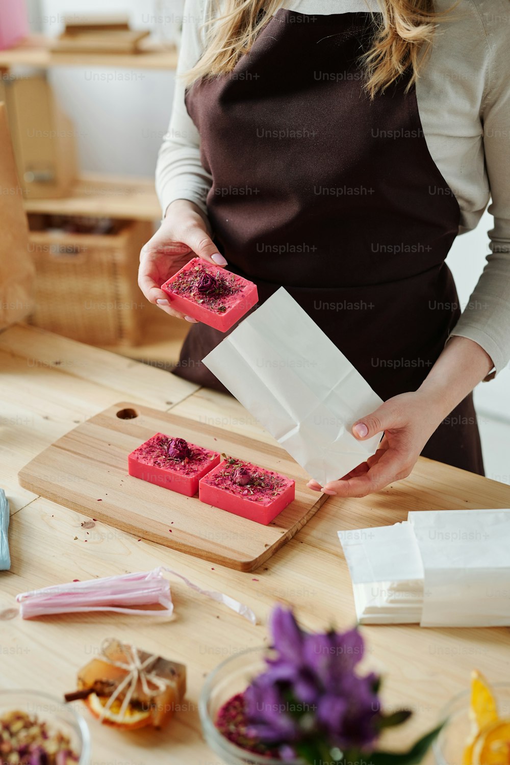 Hands of woman putting handmade pink soap bar into white paper packet over wooden table in workshop