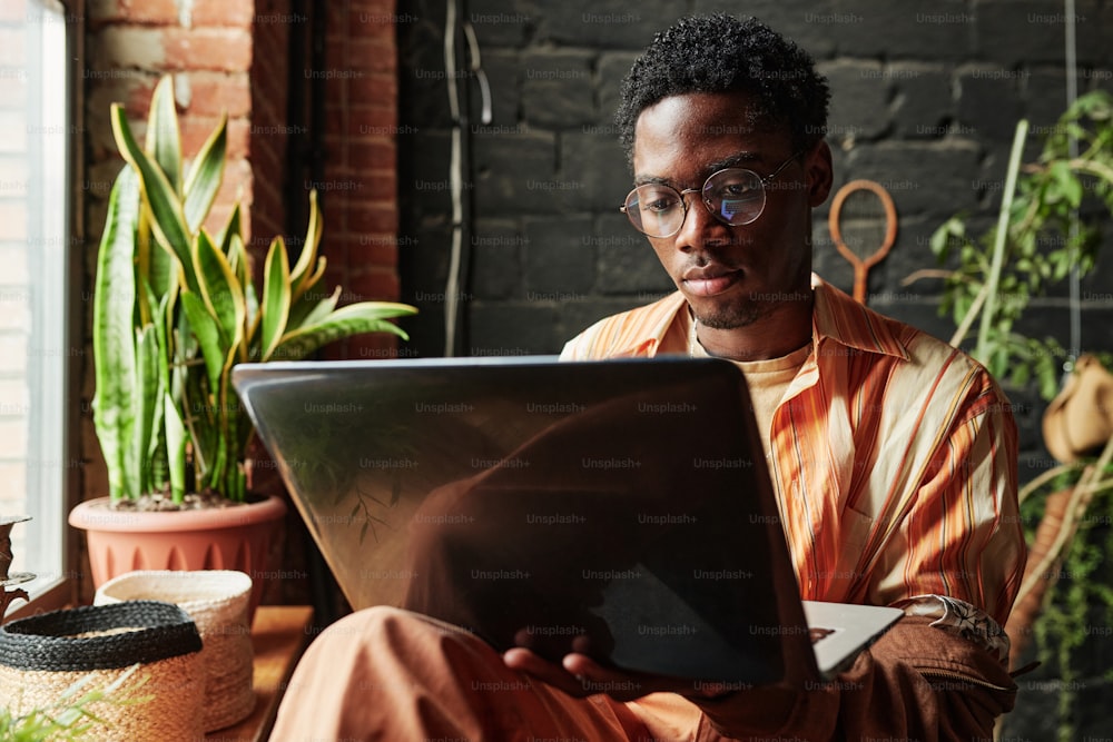 Giovane uomo nero serio in abbigliamento casual e occhiali da vista usando il computer portatile mentre è seduto vicino alla finestra nel soggiorno di un moderno appartamento loft