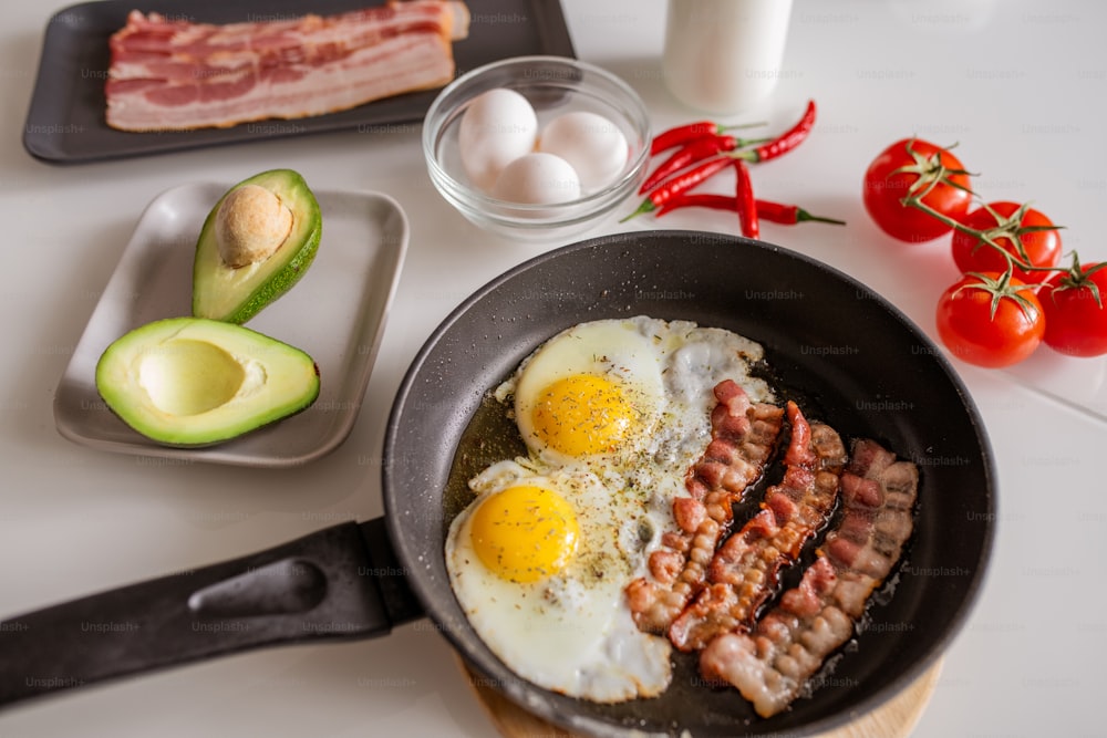 Frying pan with fried eggs and bacon sprinkled with spices, fresh avocado, red ripe tomatoes and hot chili pepper on served kitchen table