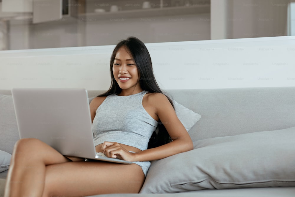 Schöne Frau mit Computer auf der Couch im Wohnzimmer zu Hause am Morgen. Glückliches lächelndes asiatisches Mädchen mit Laptop entspannt sich auf dem Sofa drinnen