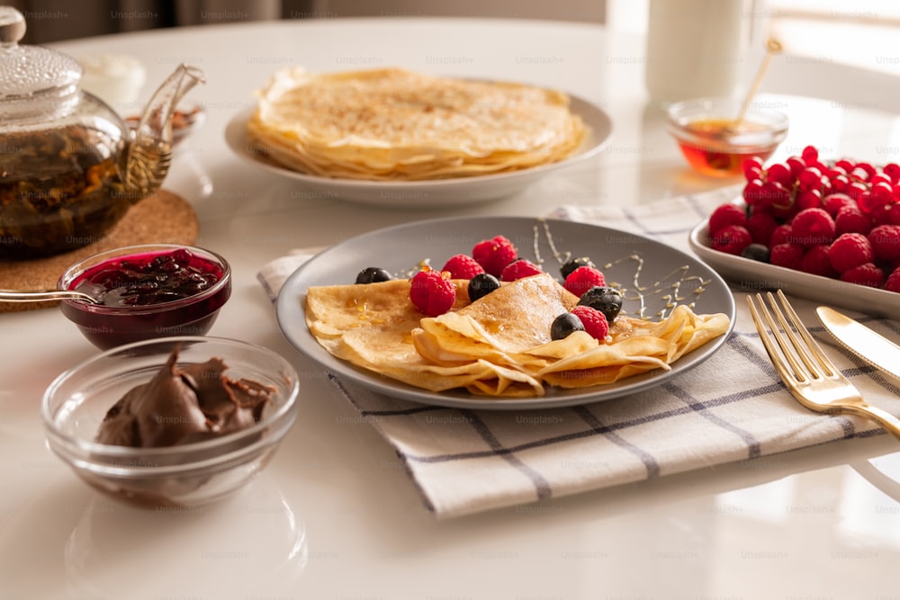 Fresh homemade appetizing crepes with berries and honey on plate, bowls with cherry jam and chocolate cream, teapot and ripe raspberries