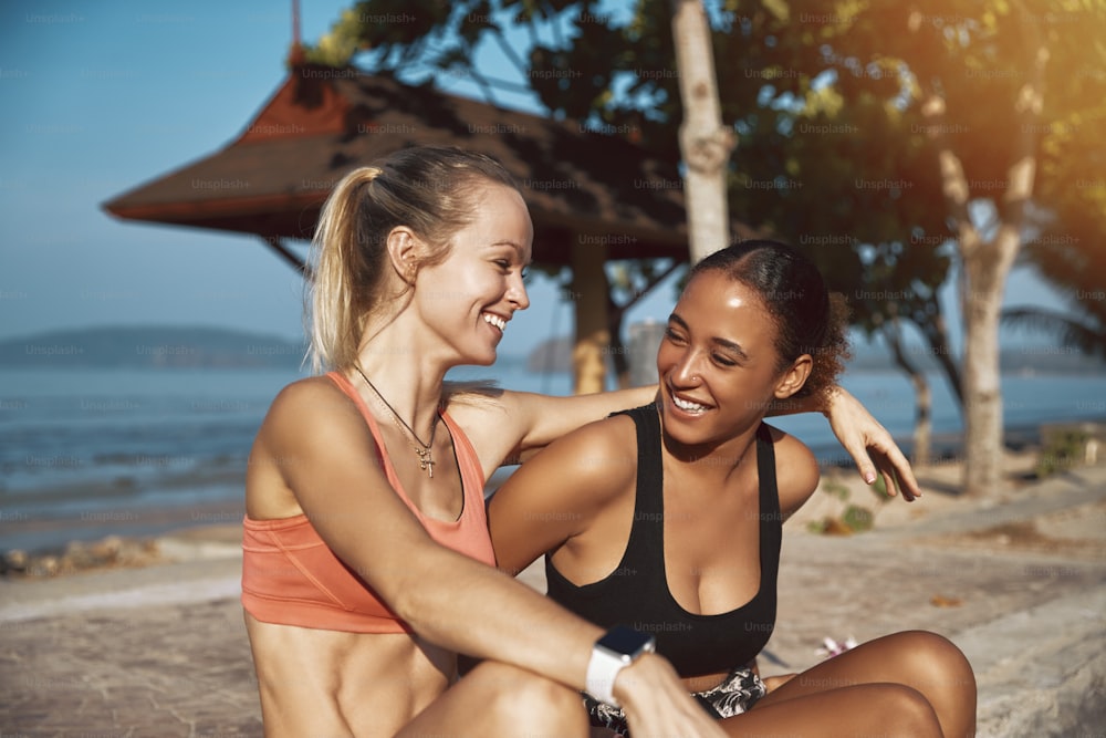 Dos mujeres jóvenes en forma en ropa deportiva riendo mientras están sentadas en una acera tomando un descanso de su trote junto al océano