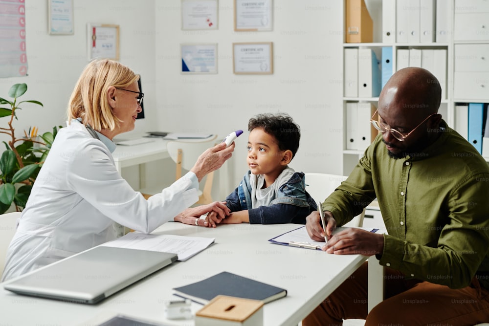 Mature female pediatrician measuring body temperature of little patient with electronic thermometer while young man filling in medical form