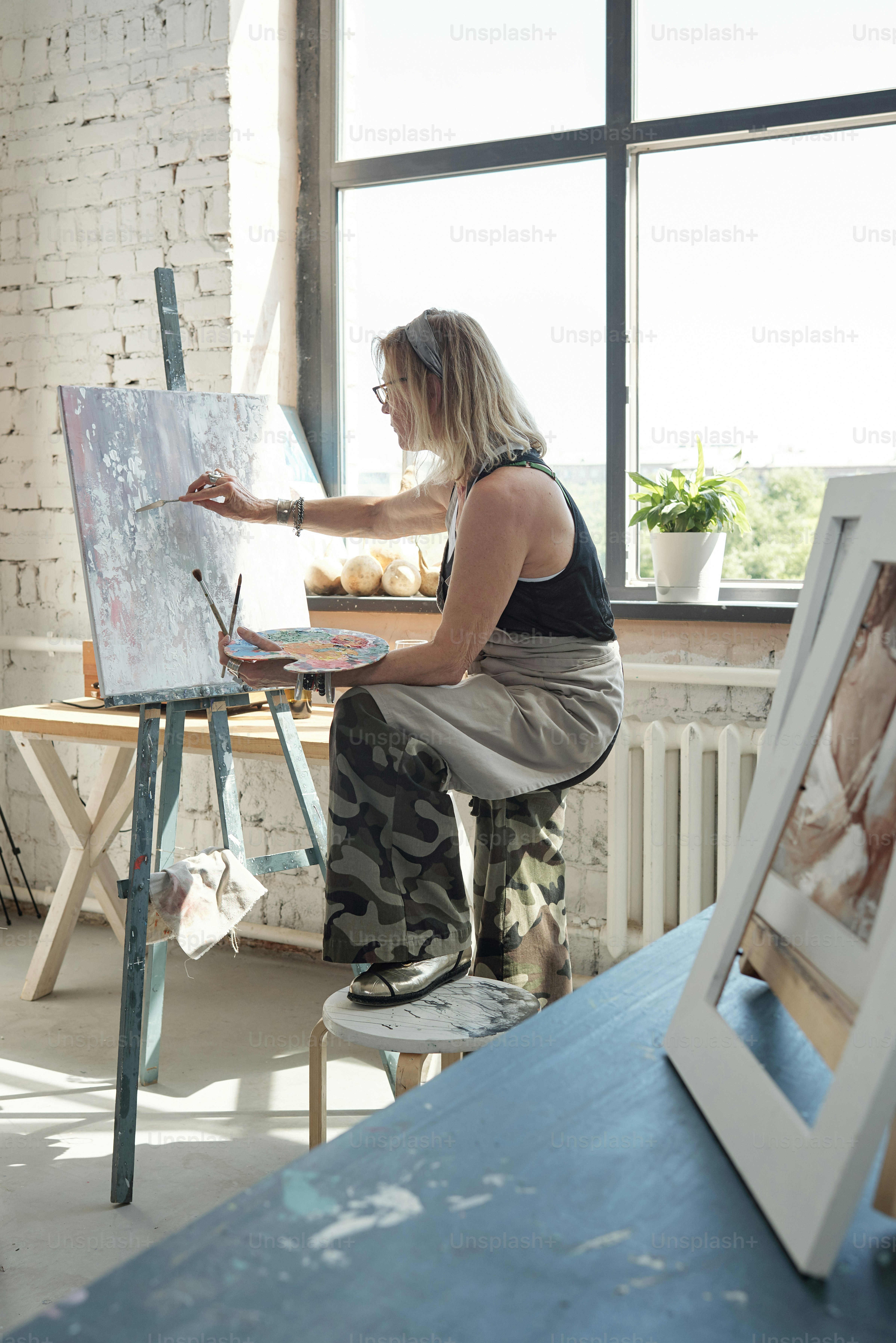 Modern blond-haired artist in glasses holding foot on stool and painting on canvas using palette