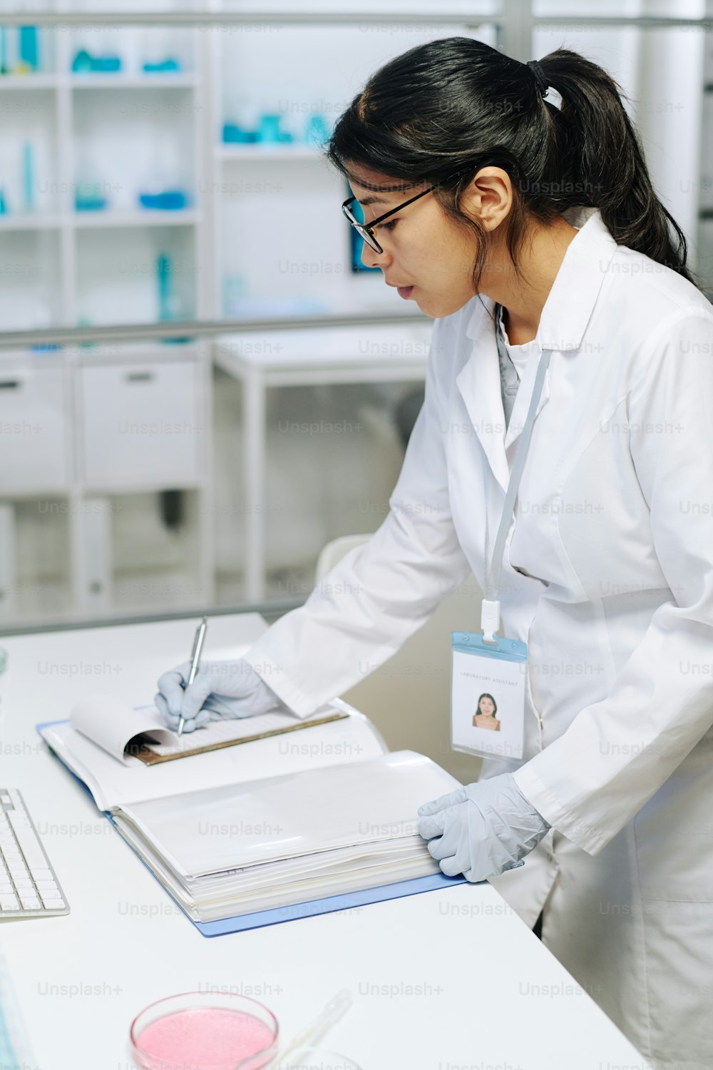 Jeune femme biochimiste en blouse blanche et gants faisant des notes scientifiques dans le bloc-notes tout en se tenant près du bureau en laboratoire clinique