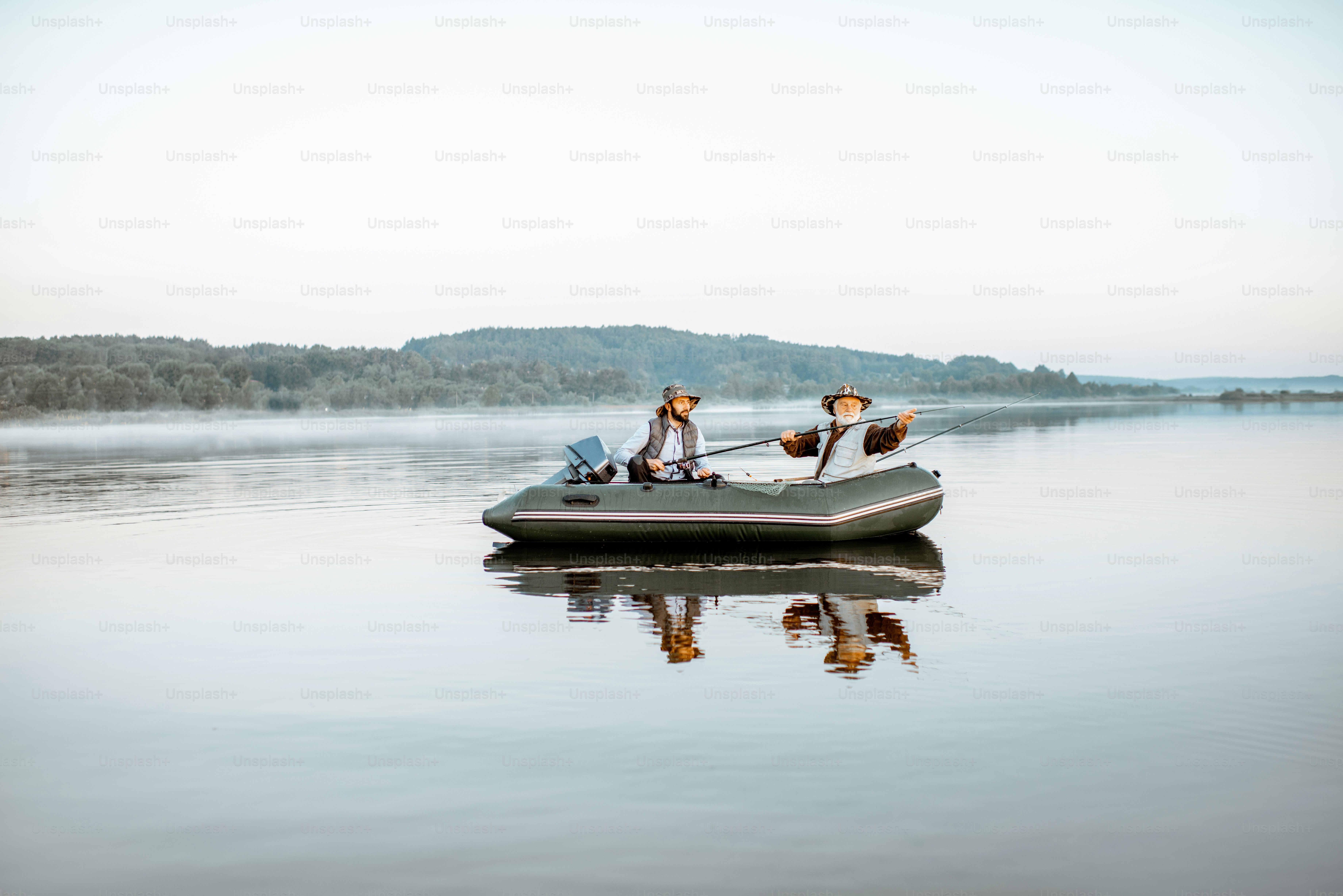 Avô com filho adulto pescando no barco inflável no lago com água calma no início da manhã. Ampla vista panorâmica