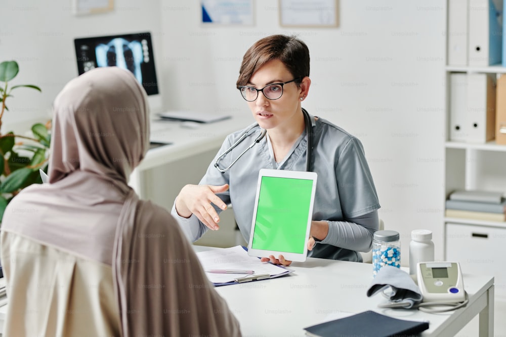 Jeune travailleur de la santé confiant tenant une tablette lors d’une présentation médicale à une patiente musulmane portant le hijab dans les cliniques