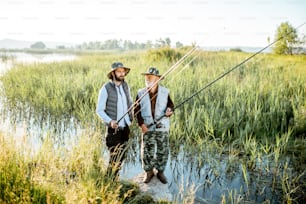 Grandfather with adult son fishing on the lake, standing together between the reeds during the morning light