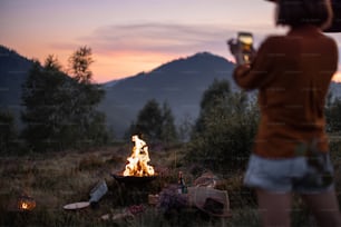 Woman photographing a bonfire on phone, having a beautiful picnic in the mountains. Travel lifestyle use of mobile devices in nature
