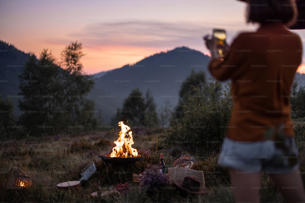 Donna che fotografa un falò sul telefono, facendo un bel picnic in montagna. Stile di vita in viaggio: uso di dispositivi mobili in natura
